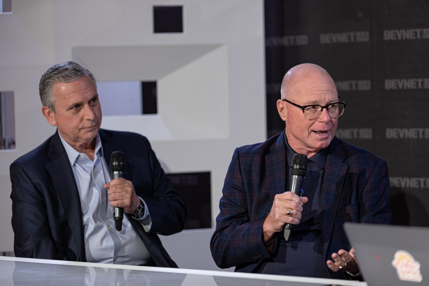 Two men in dark jackets, one with glasses and one with gray hair, hold microphones at a white desk with a laptop.