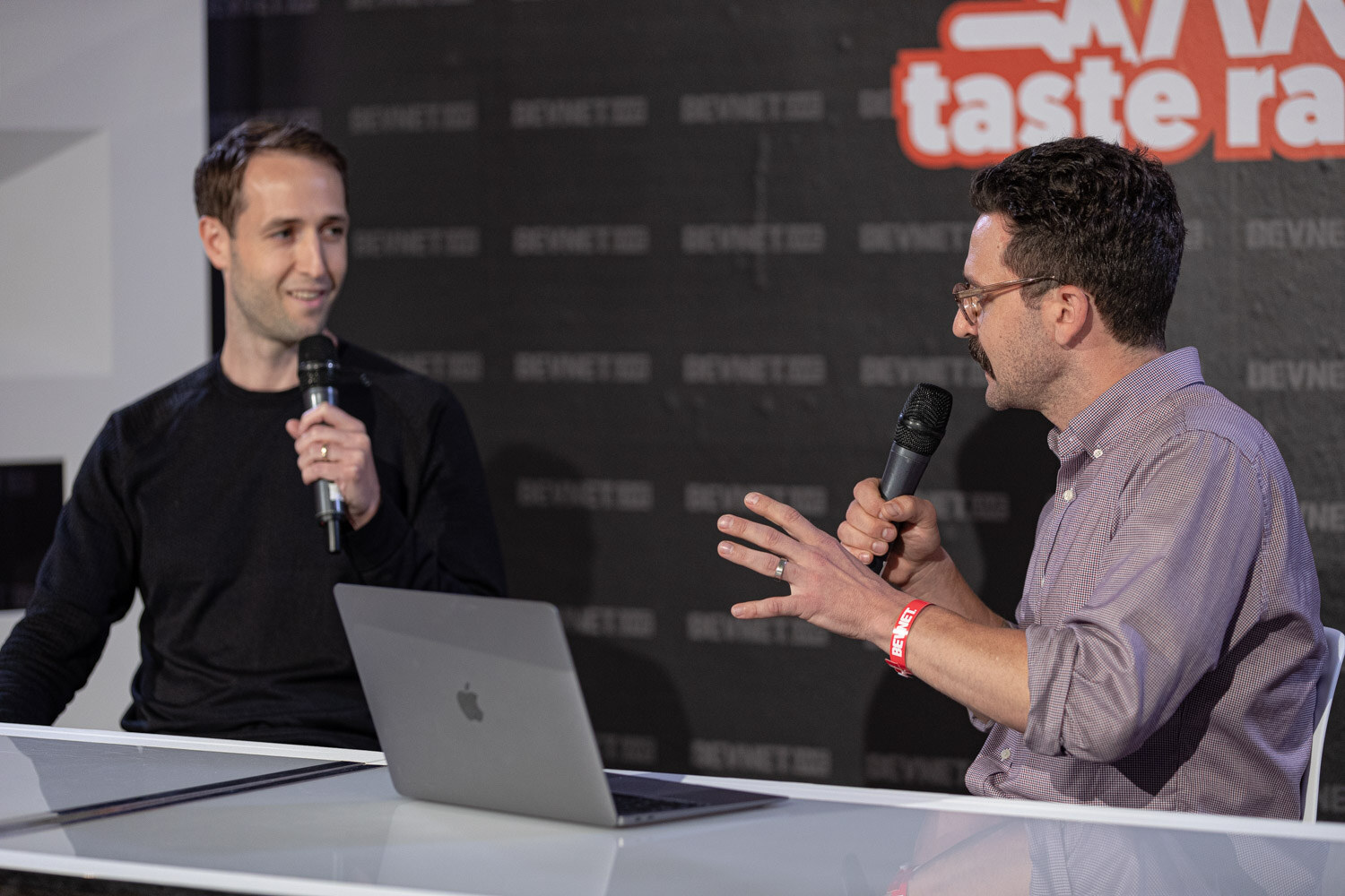 A man in a black sweater holds a microphone and smiles while a mustached man in a lavender shirt talks to him.