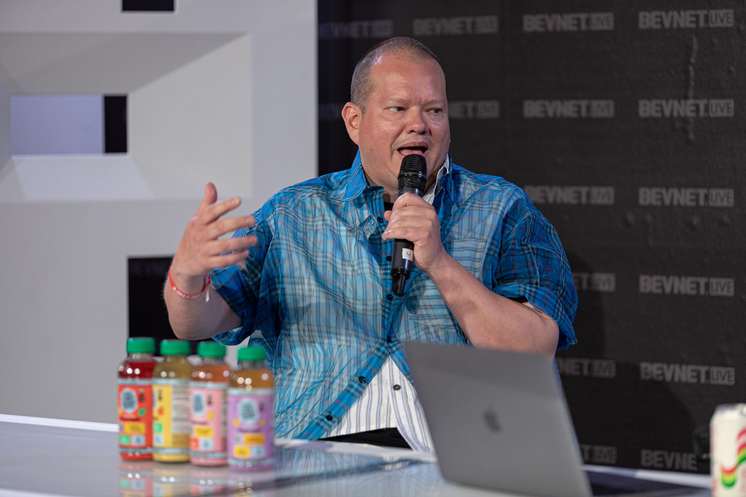 A man in a blue plaid shirt gestures while speaking into a microphone with four colorful bottles of drinks on the desk in front of him.