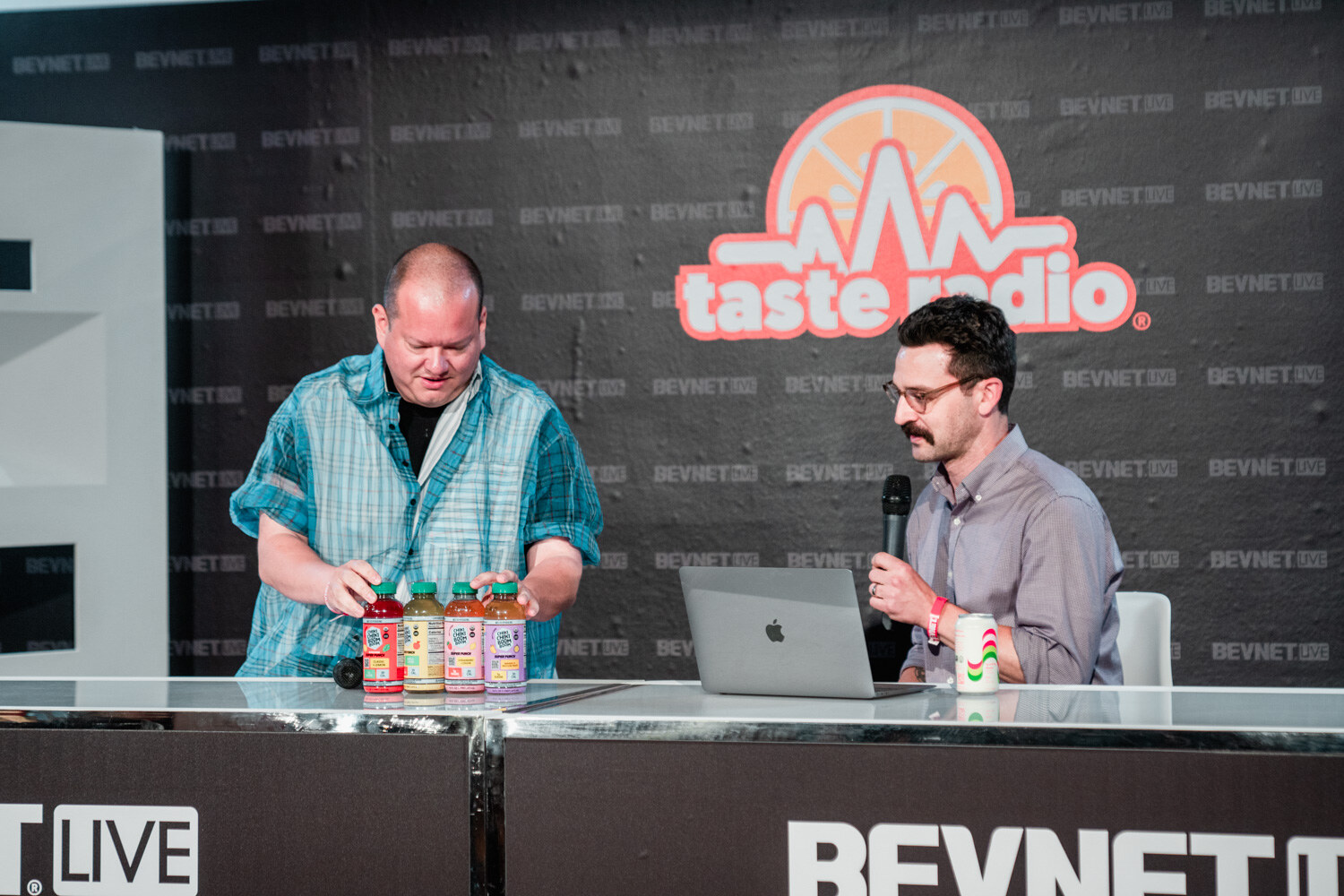 A man in a blue plaid shirt arranges bottles on the desk as a man in a gray shirt with a mustache holds a microphone.