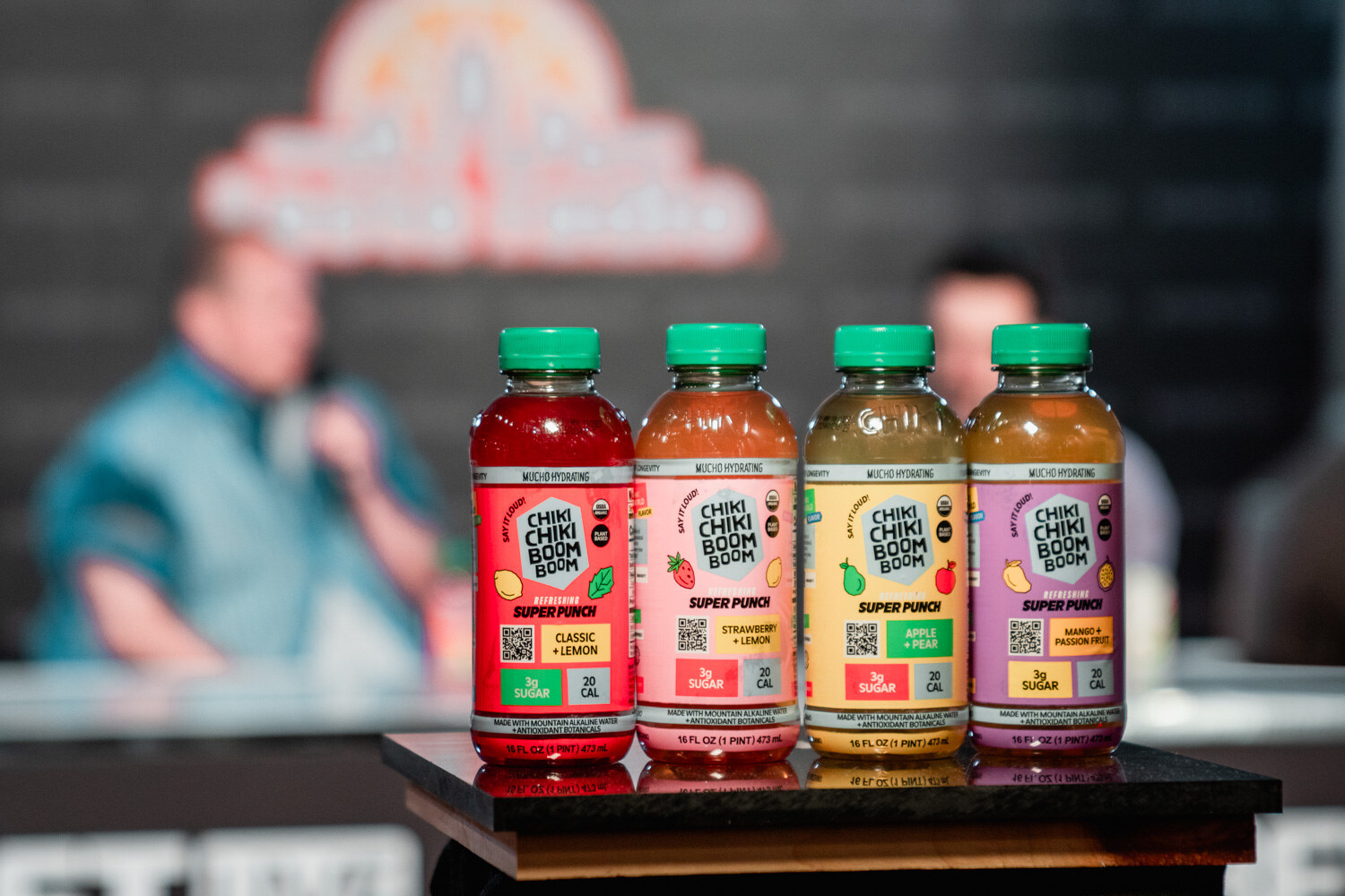 Four bottles labeled Chiki Chiki Boom Boom Super Punch in red, orange, yellow and purple, with two blurred people behind.