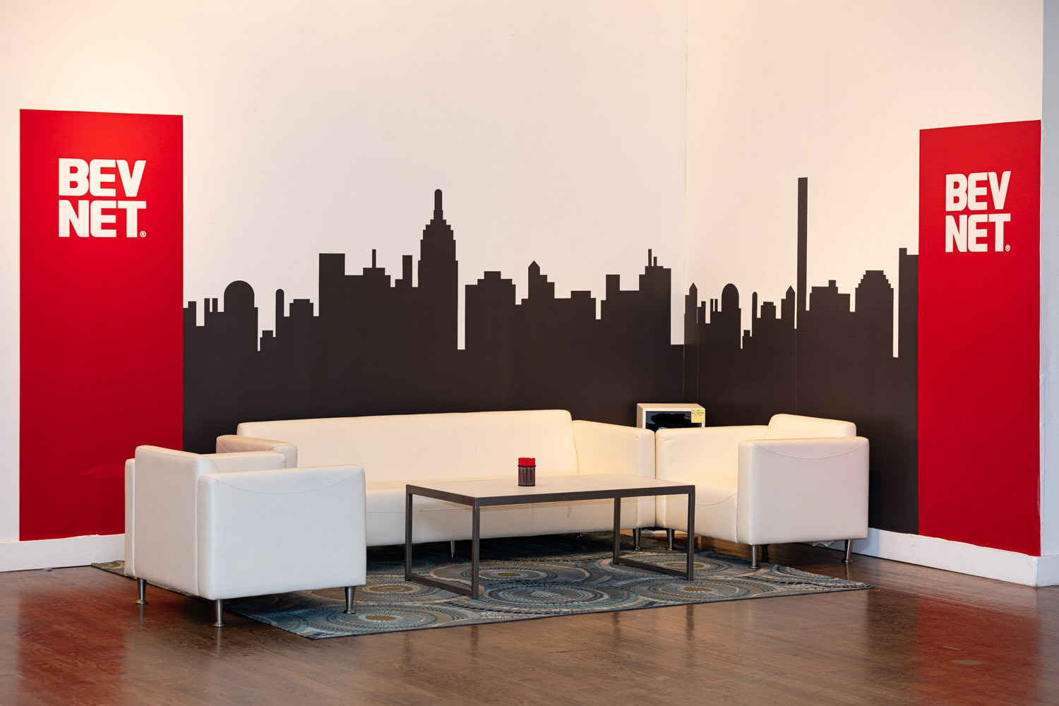 A seating area with white couches and a coffee table in front of a black skyline silhouette mural, two red BevNET banners on either side.