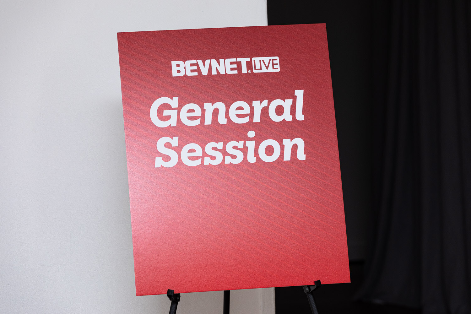 A red easel sign reading BevNET Live General Session in white text, in front of a black curtain.