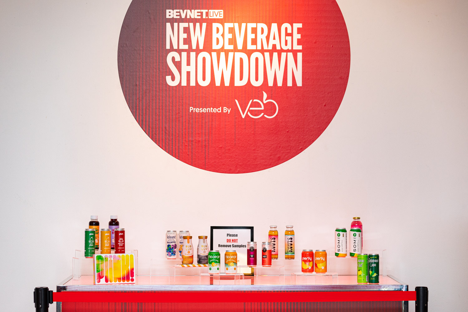 A table with rows of bottles and cans of drinks beneath a round sign reading BevNET Live New Beverage Showdown Presented by Veb.