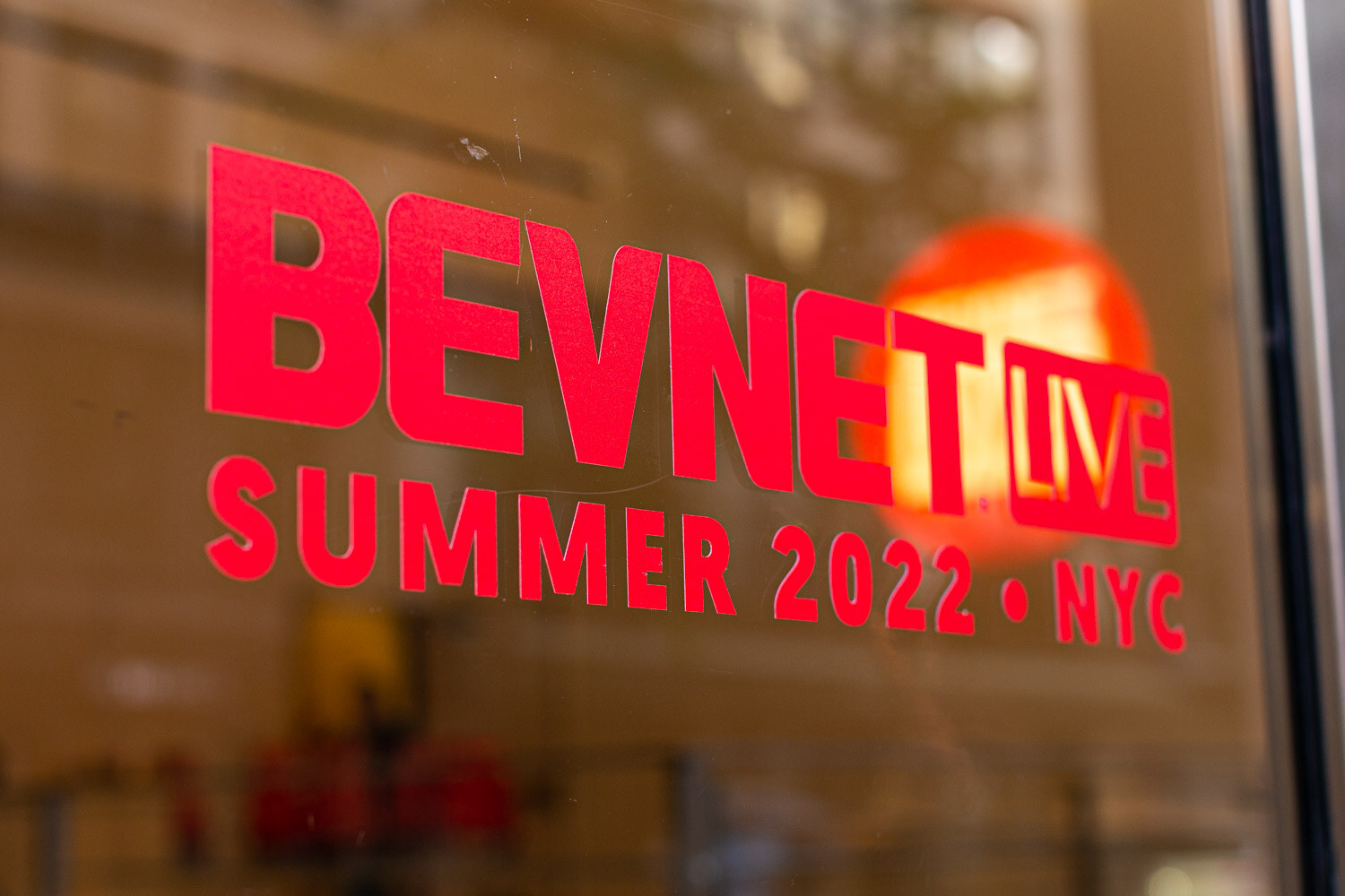 A red decal reading BevNET Live Summer 2022 NYC on a glass window with reflections behind it.