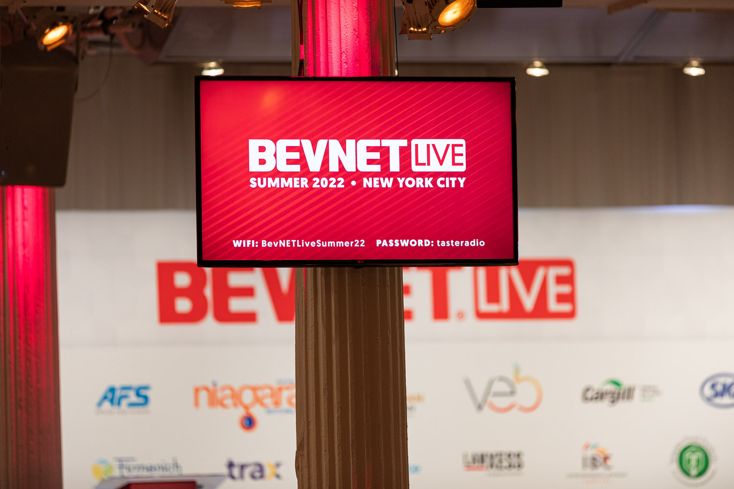 A red monitor on a pole reads BevNET Live Summer 2022 New York City, with the BevNET Live stage backdrop blurred behind.