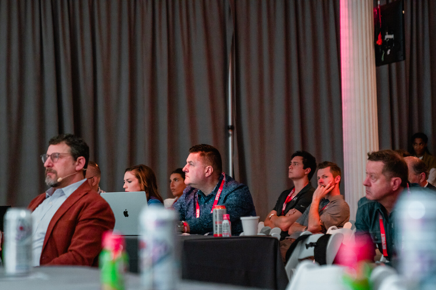 A row of seated attendees in lanyards, some with laptops, look toward the stage with drink cans in the foreground.