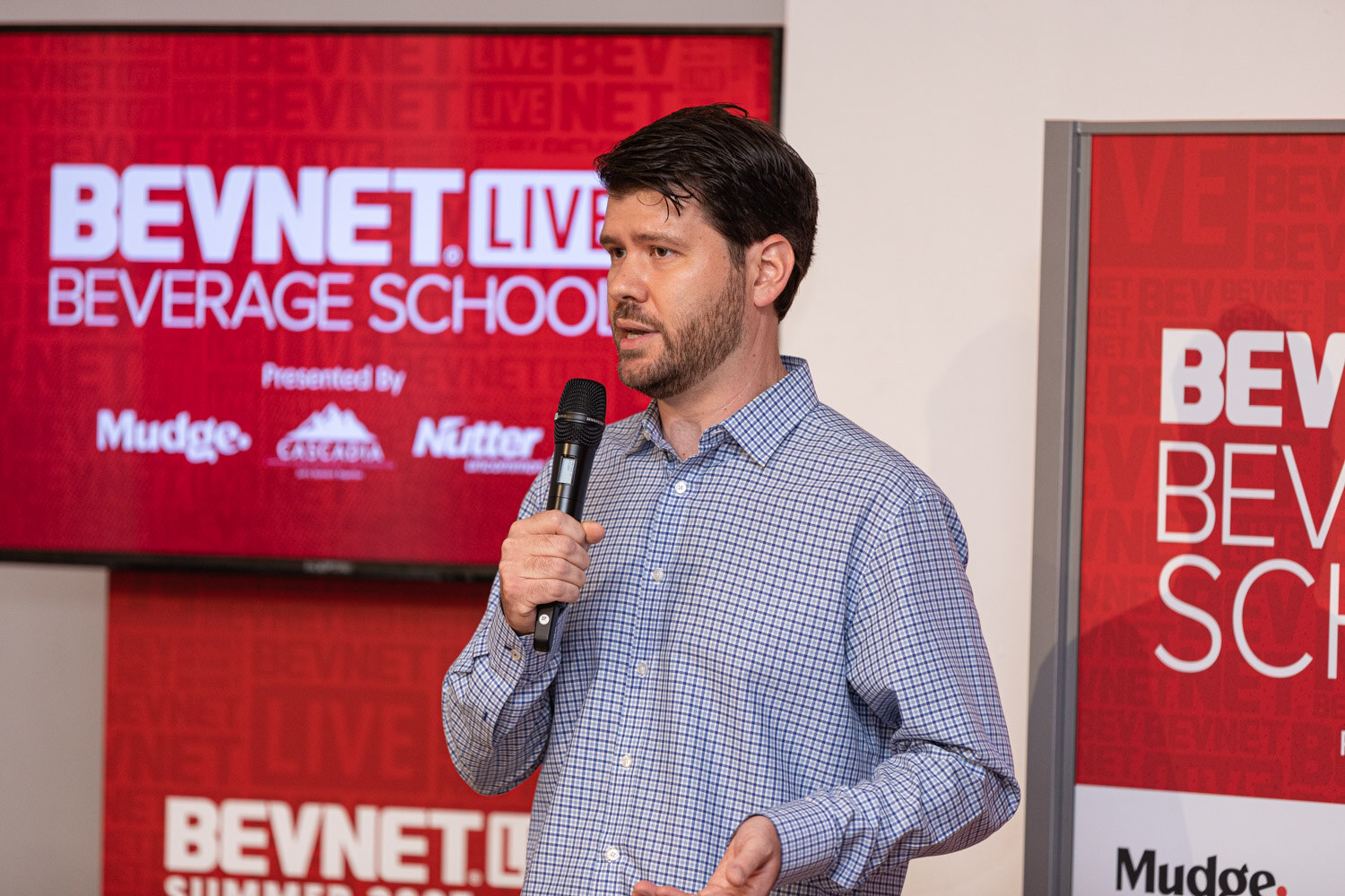 A man in a blue checked shirt speaks into a microphone beside a red screen reading BevNET Live Beverage School with sponsor logos including Mudge.