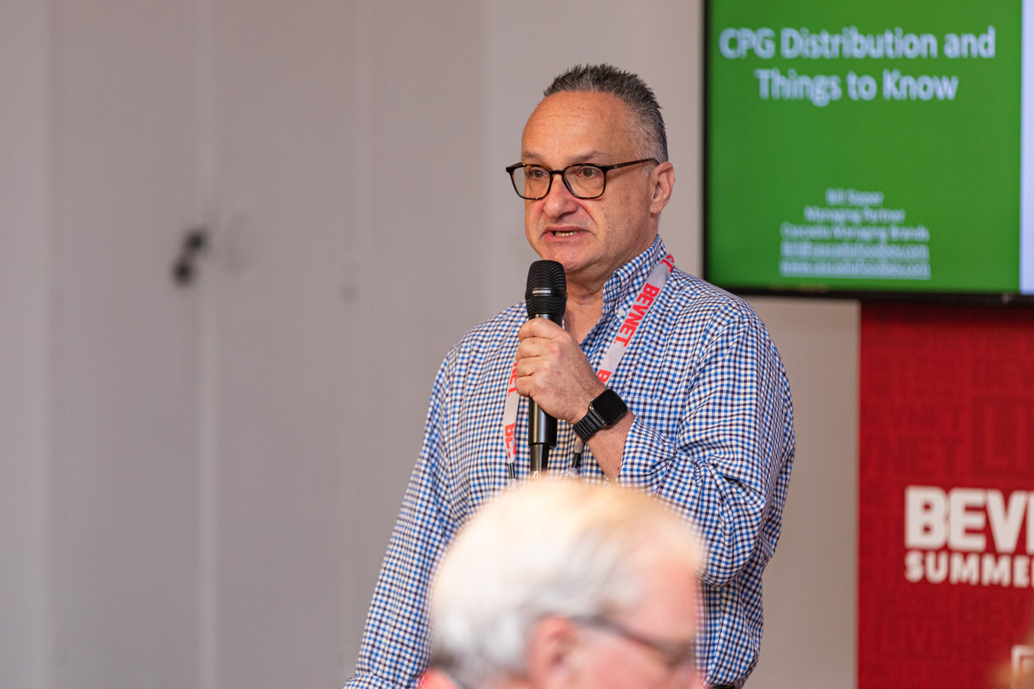 A man with glasses in a blue checked shirt and red lanyard holds a microphone beside a screen titled CPG Distribution and Things to Know.