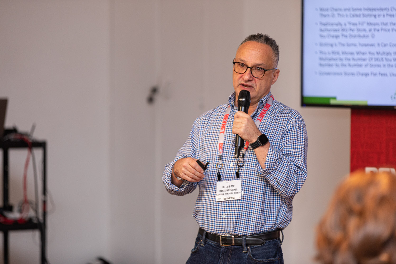 A man with glasses in a checked shirt and red lanyard holds a microphone and gestures in front of a slide and a BevNET banner.