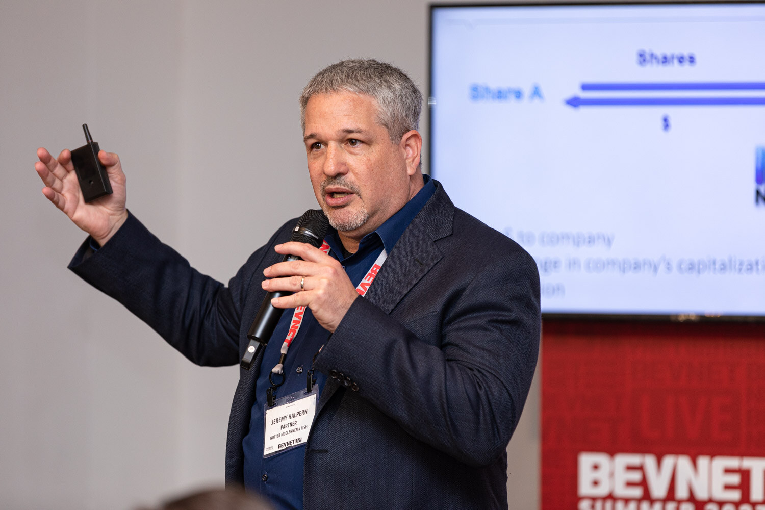 A gray-haired man in a navy blazer holds a clicker in one hand and a microphone in the other in front of a slide with a labeled diagram.