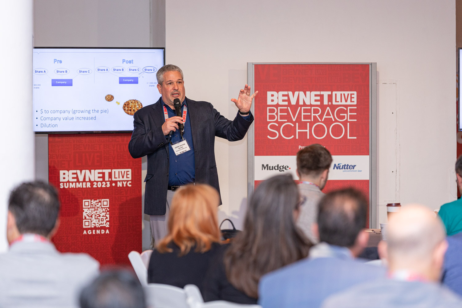 A man in a navy blazer gestures while speaking in front of a red BevNET Live Beverage School sign, with seated attendees in the foreground.