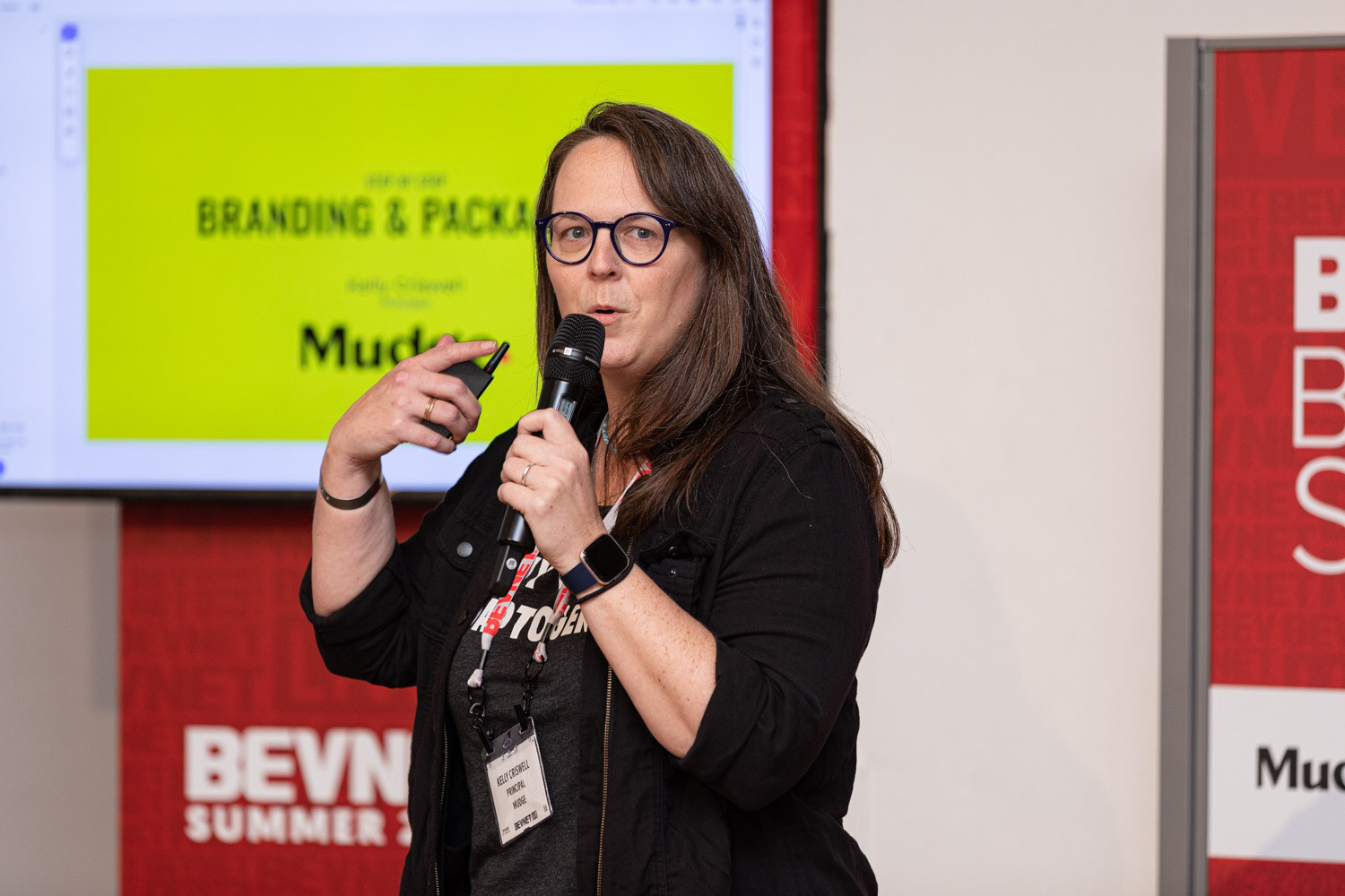 A woman with glasses and a lanyard holds a microphone and a clicker in front of a lime-green slide reading Branding and Packaging with a Mudge logo.