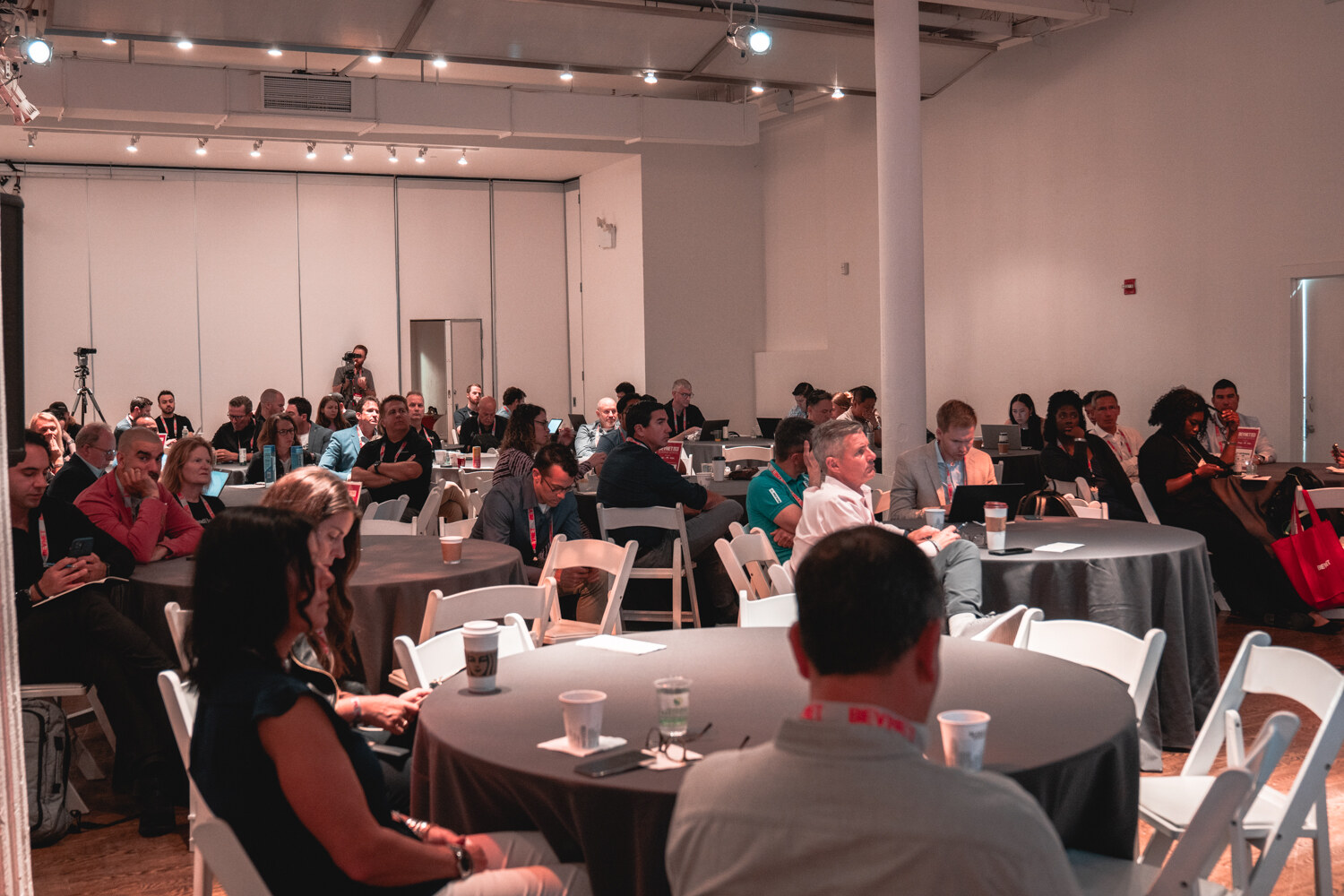 Wide view of people seated at round gray tables and white folding chairs in a loft room with track lighting.