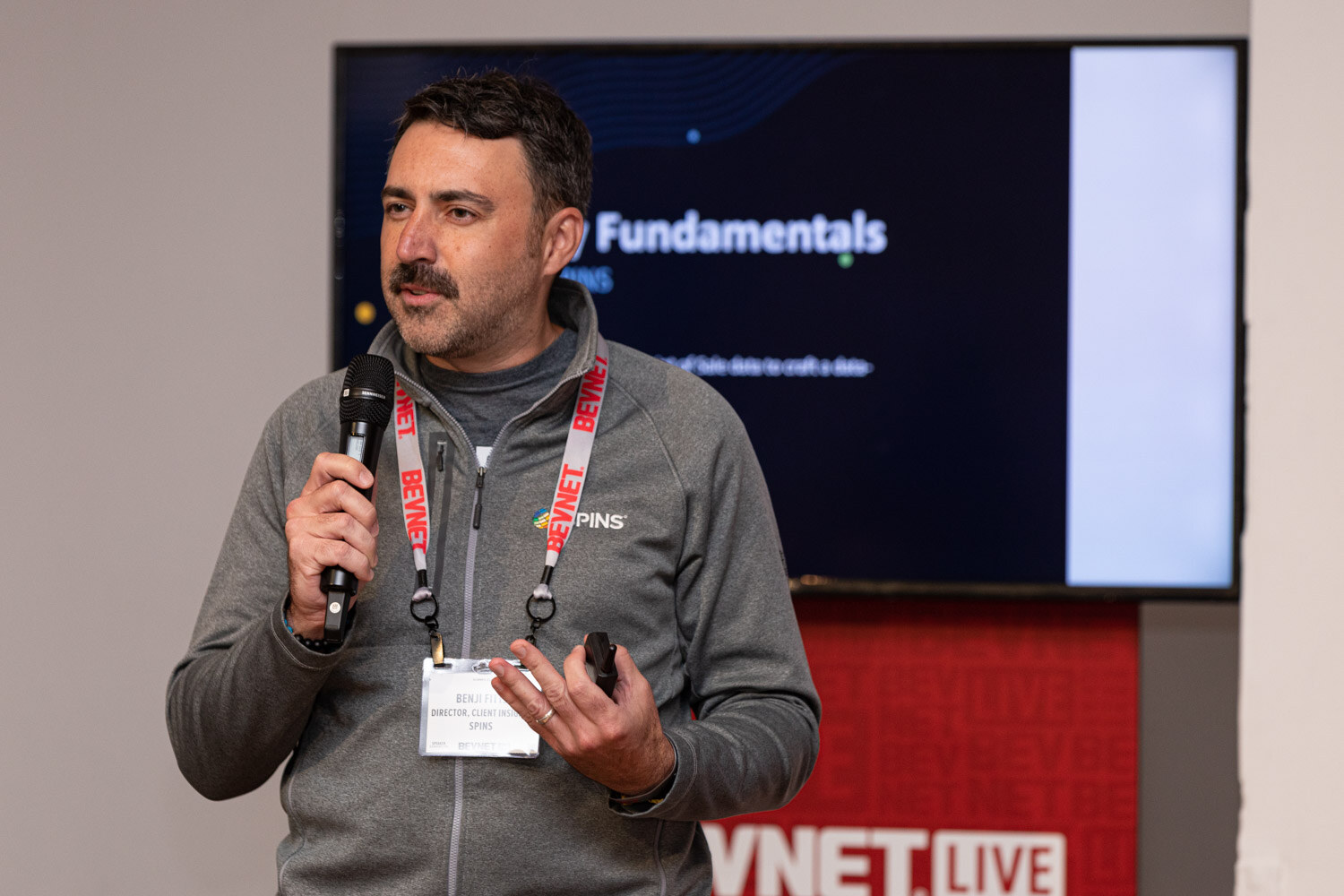 A man with a mustache in a gray pullover and lanyard holds a microphone in front of a screen with a slide reading Fundamentals.
