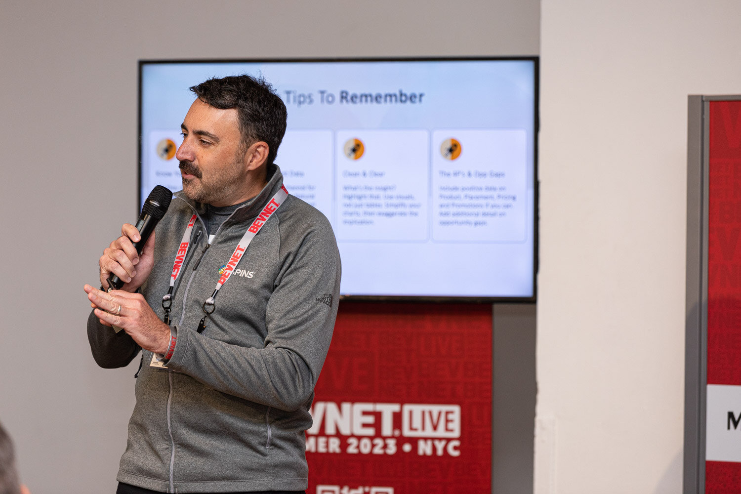 A man in a gray pullover with a lanyard holds a microphone in front of a screen with a slide titled Tips To Remember and a BevNET Live Summer 2023 banner.