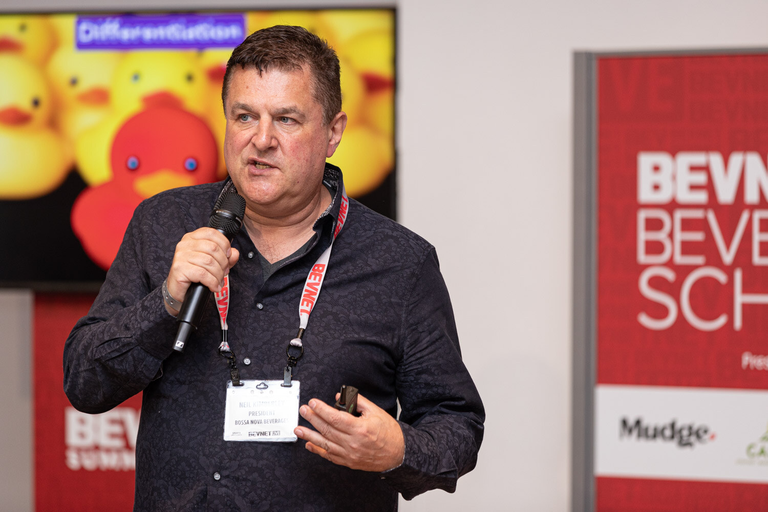 A man in a dark button-up shirt and red lanyard holds a microphone in front of a screen showing rubber ducks and the word Differentiation.