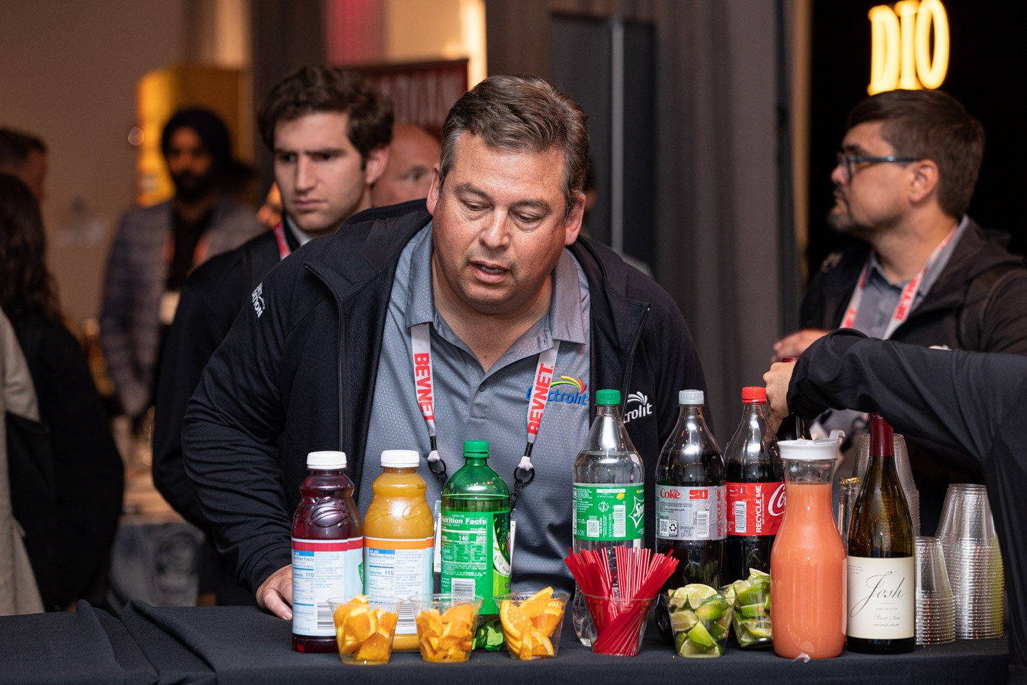 A man with a lanyard leans over a black table with bottles of juice and soda, cut fruit and red straws, with a DIO sign glowing behind him.