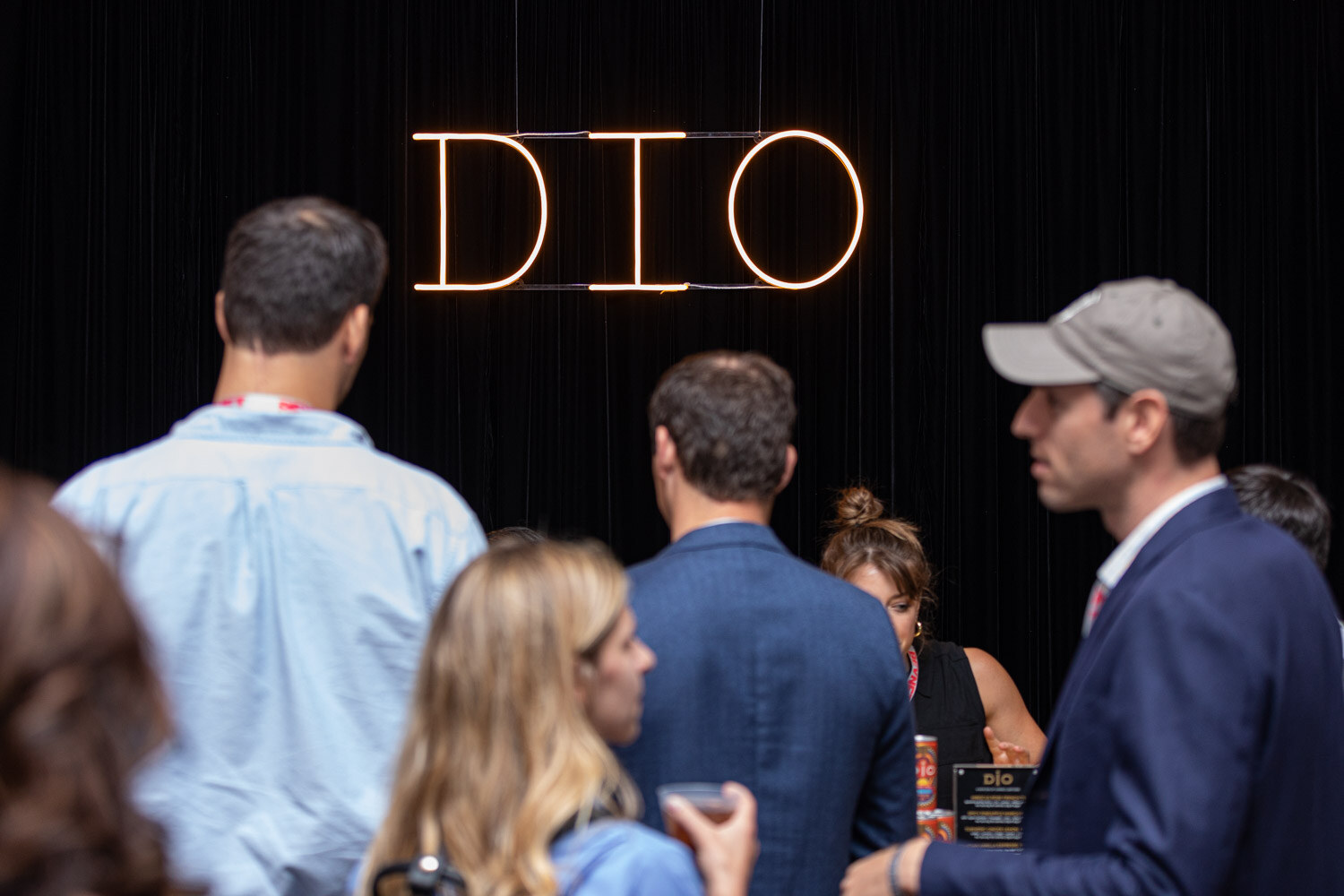 Several attendees stand in front of a dark curtain with a glowing DIO sign while a bartender works behind them.
