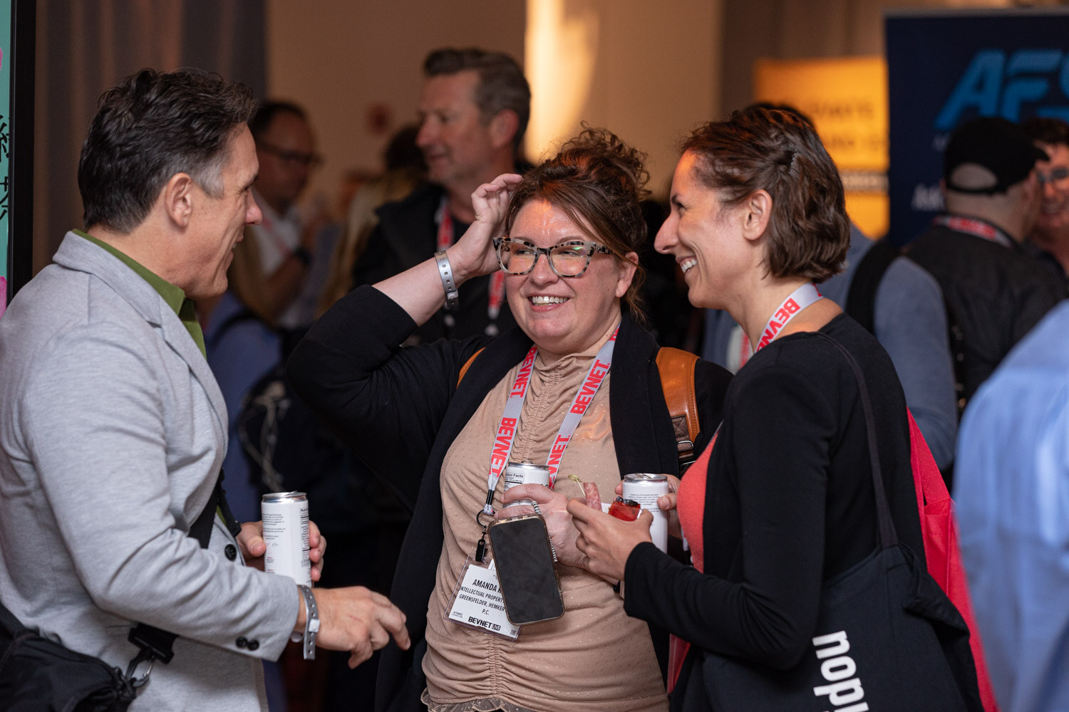 A group of attendees with lanyards laugh and talk in a crowded room, one woman with glasses holding a phone and a can.