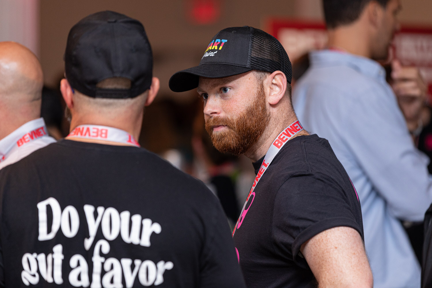 A man in a black cap with a Do your gut a favor shirt and a bearded man in a Hart Print cap with a lanyard talk in a busy room.