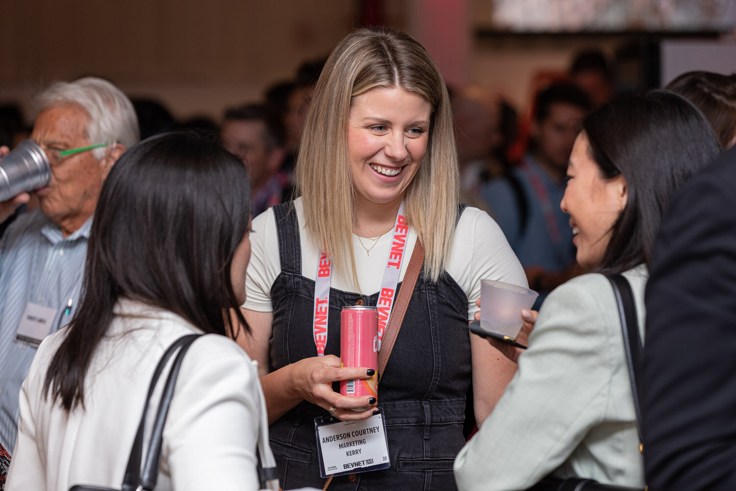 A smiling blonde woman in a black overall dress holds a pink can and talks with other attendees with lanyards in a crowded room.