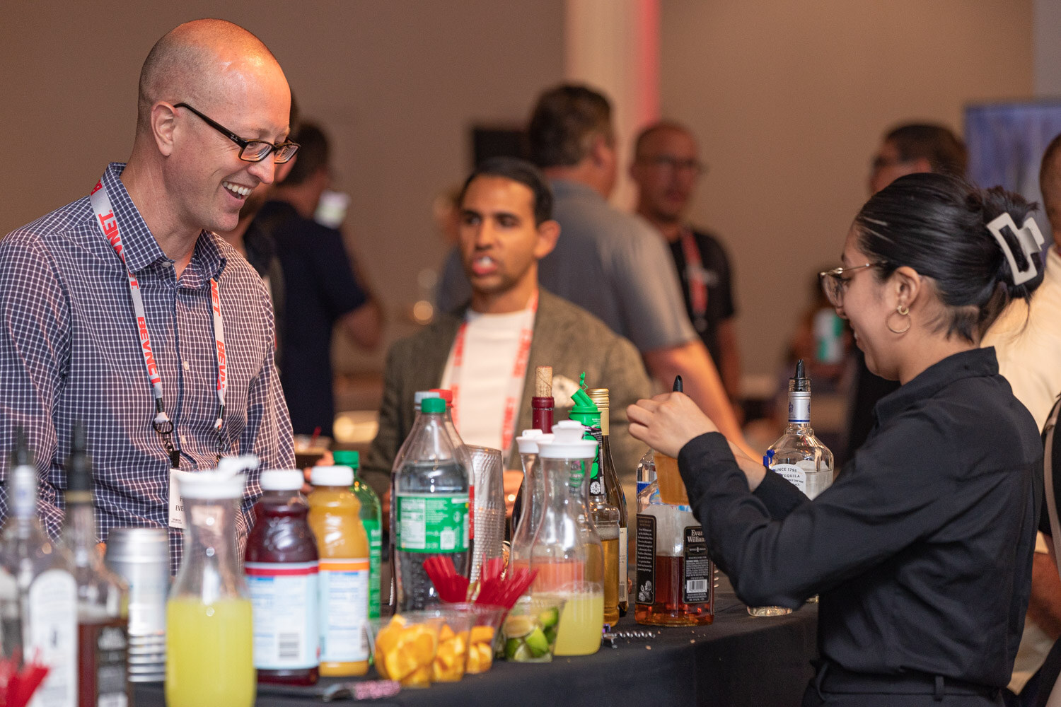 A smiling man in a checked shirt and lanyard stands at a bar with bottles of juice and cut fruit while a bartender in black mixes a drink.