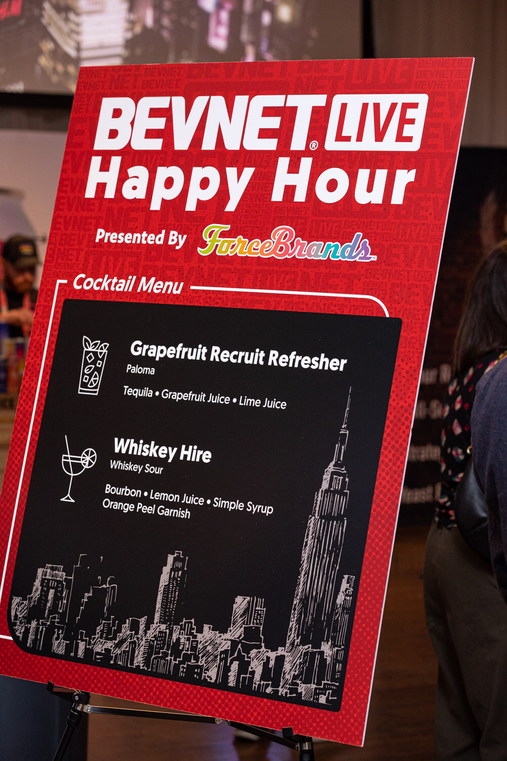 A red easel sign reading BevNET Live Happy Hour with a cocktail menu listing a Grapefruit Recruit Refresher and a Whiskey Hire, with a New York skyline drawing.