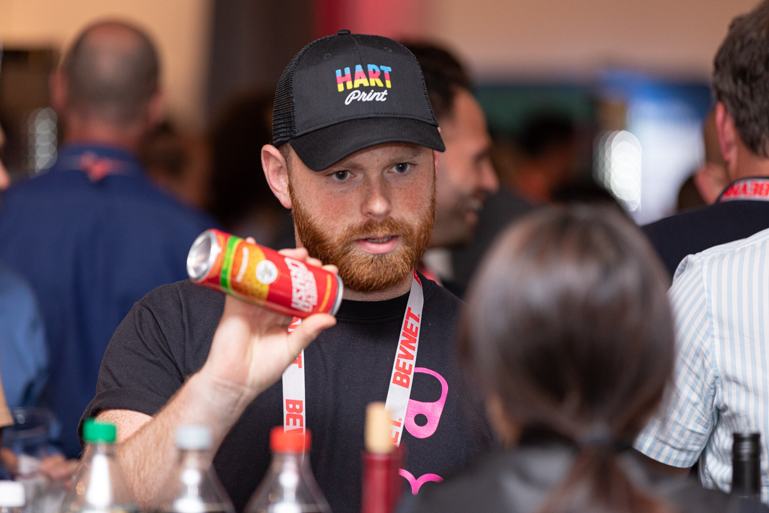 A bearded man in a black Hart Print cap and a black T-shirt with a lanyard holds up a red can while talking with another attendee.