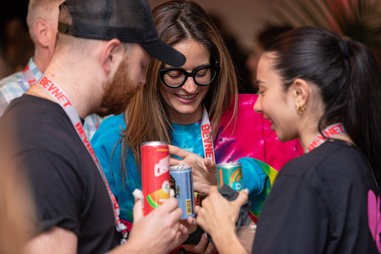 A bearded man in a cap, a woman with black glasses and a bright top, and a woman in black hold cans and look at them together.