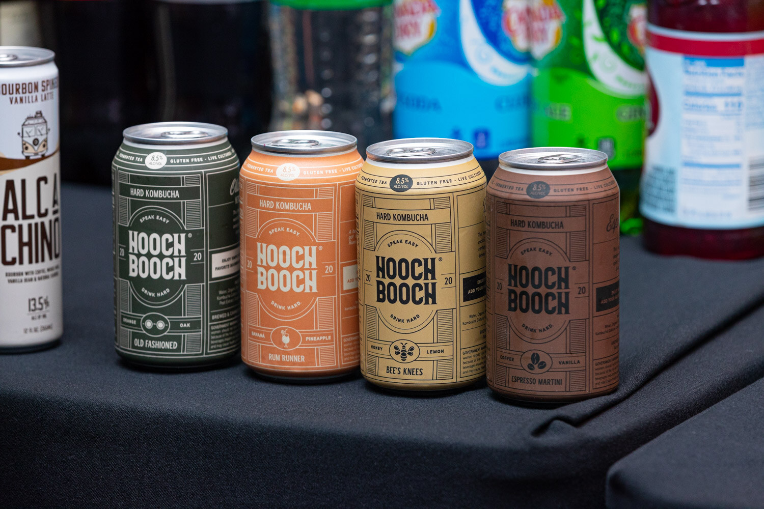 Four cans of Hooch Booch hard kombucha in green, orange, yellow and brown stand on a black tablecloth with soda and juice bottles behind them.