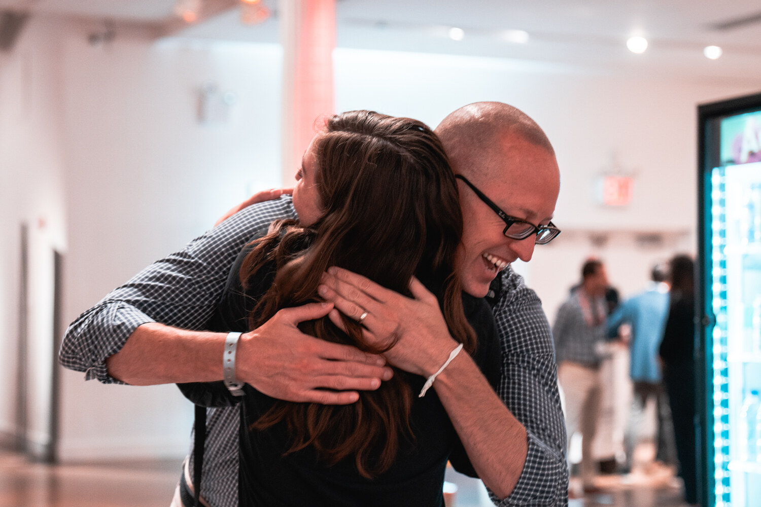 A man with glasses and a woman with long dark hair hug in a bright loft room with a lit display fridge at right.