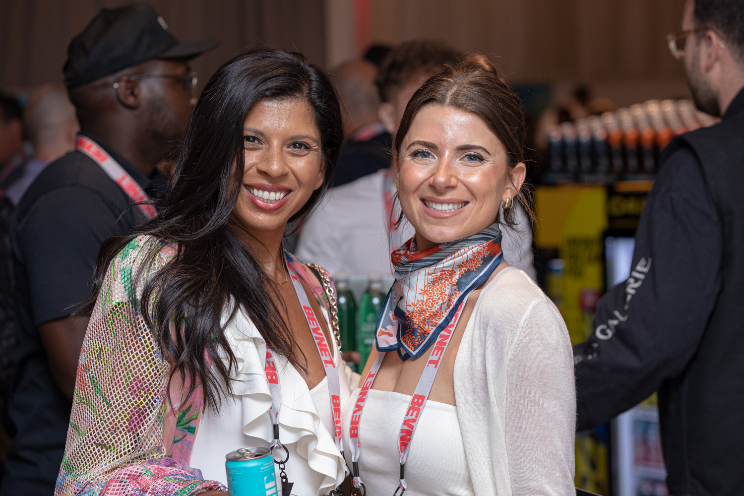 Two smiling women wearing lanyards stand close together in a crowded room, one holding a can.