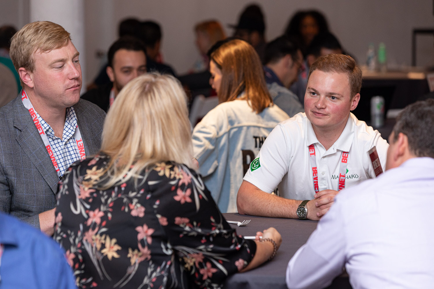 Several people with lanyards sit and talk at a round table in a busy room, one man in a white polo and others in jackets.