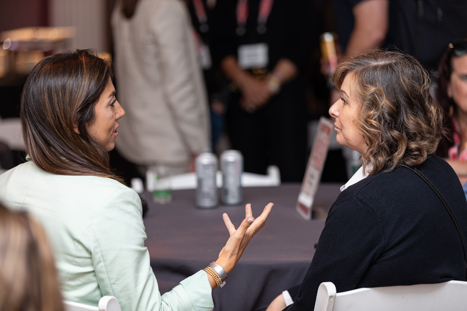 A woman in a mint jacket and a woman with wavy hair gesture while talking beside a gray tablecloth with cans on it.