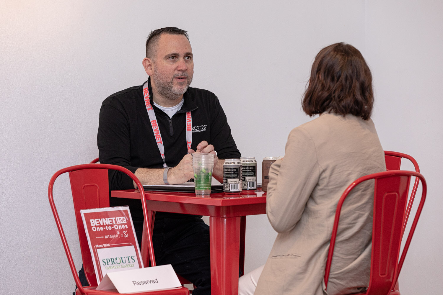 A man in a black shirt with a lanyard talks with a woman in a beige jacket at a red table with a Sprouts Reserved sign.