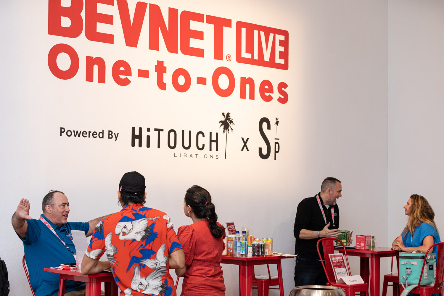 Wide view of people meeting at red high tables in front of a white wall with the BevNET Live One-to-Ones sign powered by Hi-Touch Libations.