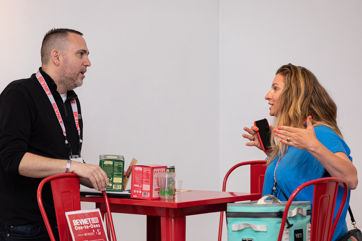 A man in a black shirt with a lanyard talks with a woman in a blue top at a red table with product cartons and a teal cooler bag.
