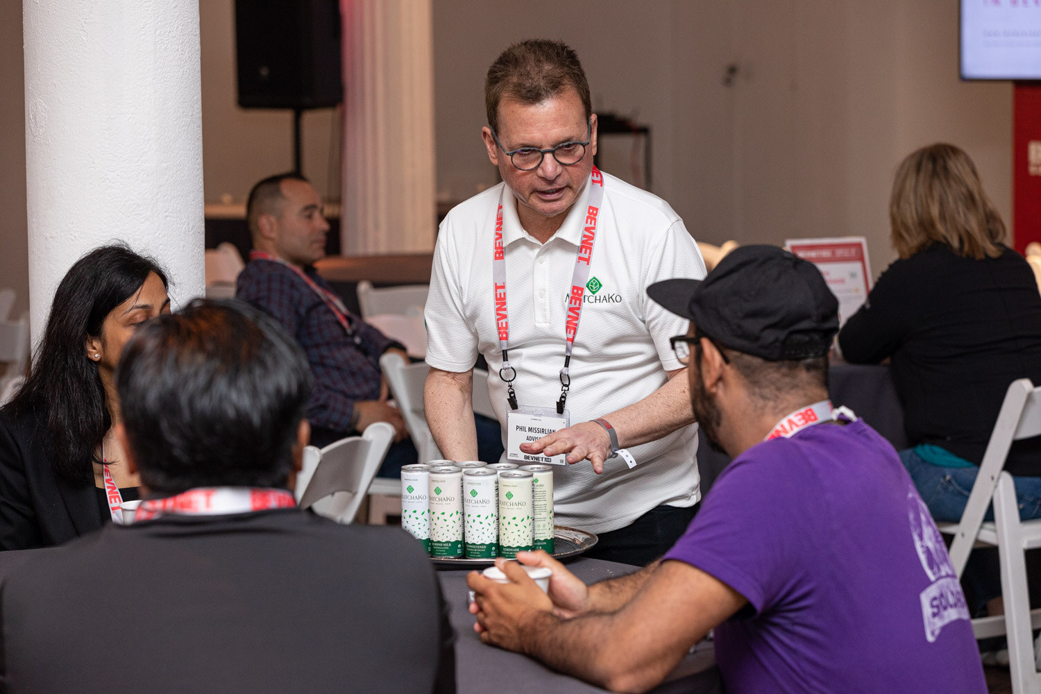 A man in a white shirt with a lanyard holds a tray of small green and white cans over a table where attendees sit, one in a purple shirt.