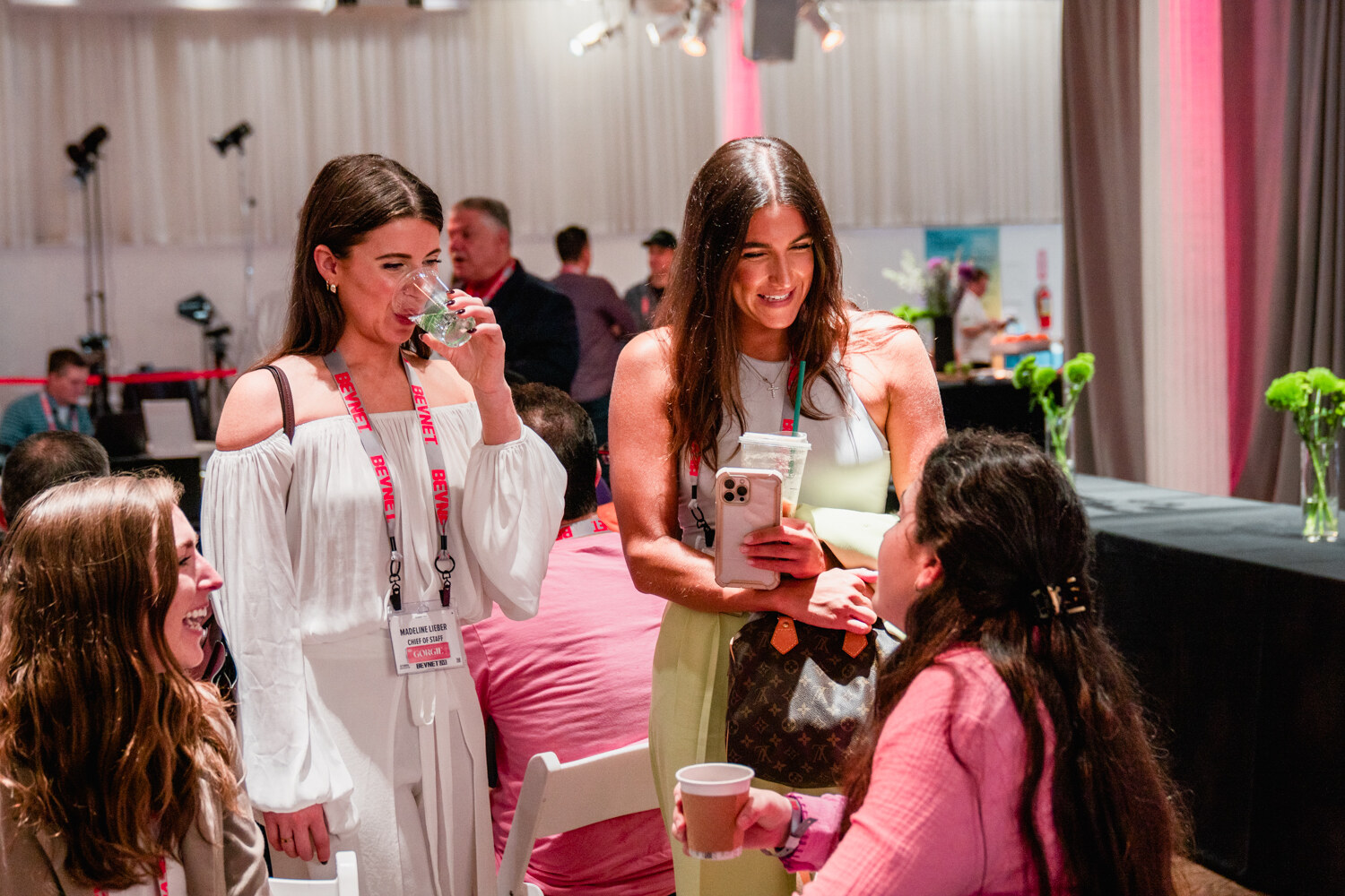 Three women with lanyards, two in white and green outfits holding drinks, talk with a woman in pink in a bright loft room.