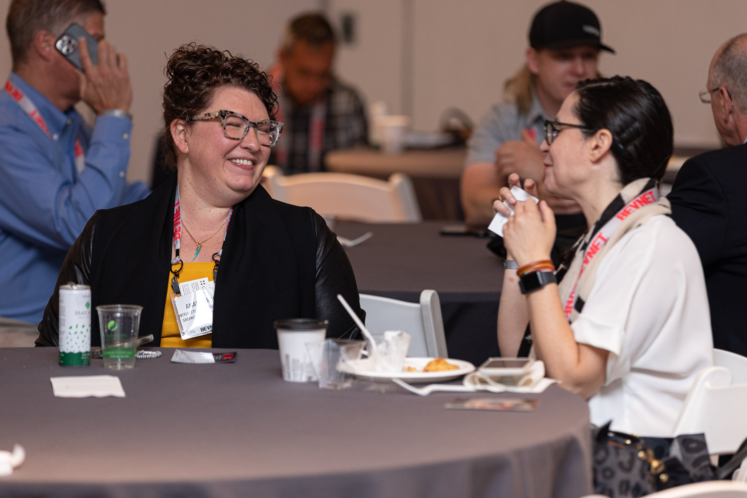 A woman with curly hair and glasses wearing a name badge laughs with a woman in a white top at a round table.