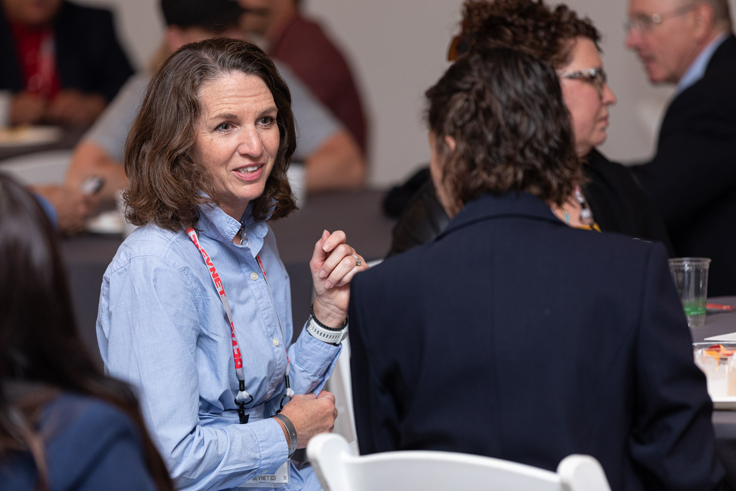 A woman with shoulder-length brown hair in a light blue button-up shirt and lanyard talks with a woman in a dark blazer at a gray table.