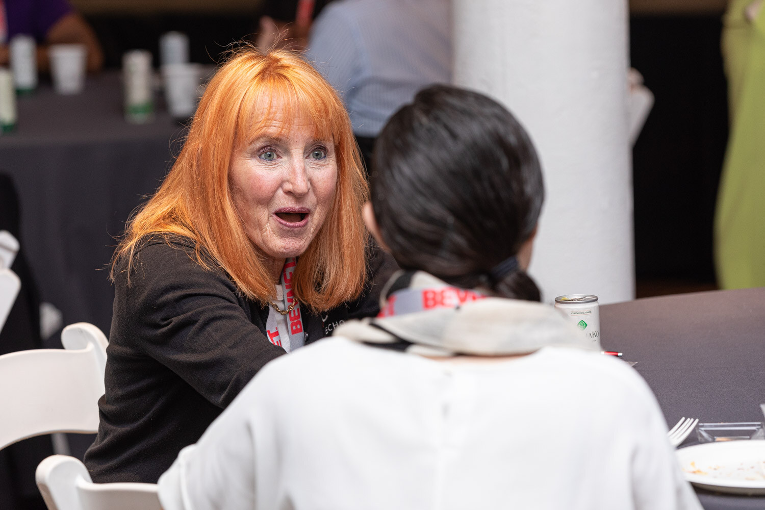 A woman with orange hair and a black jacket looks surprised while talking with a woman in a white top at a round table.