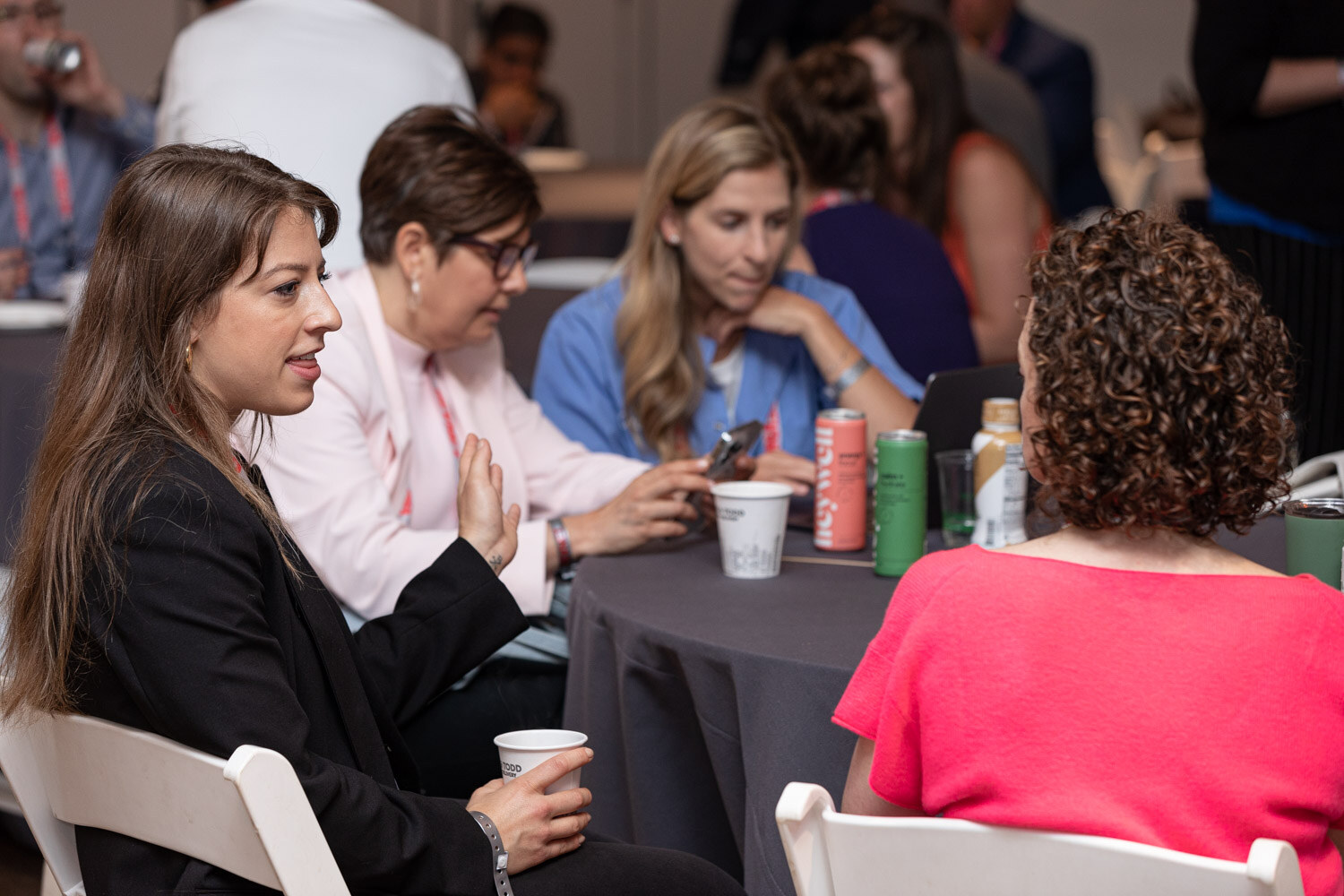 Four women sit at a round gray table talking, with cans and plastic cups in front of them.
