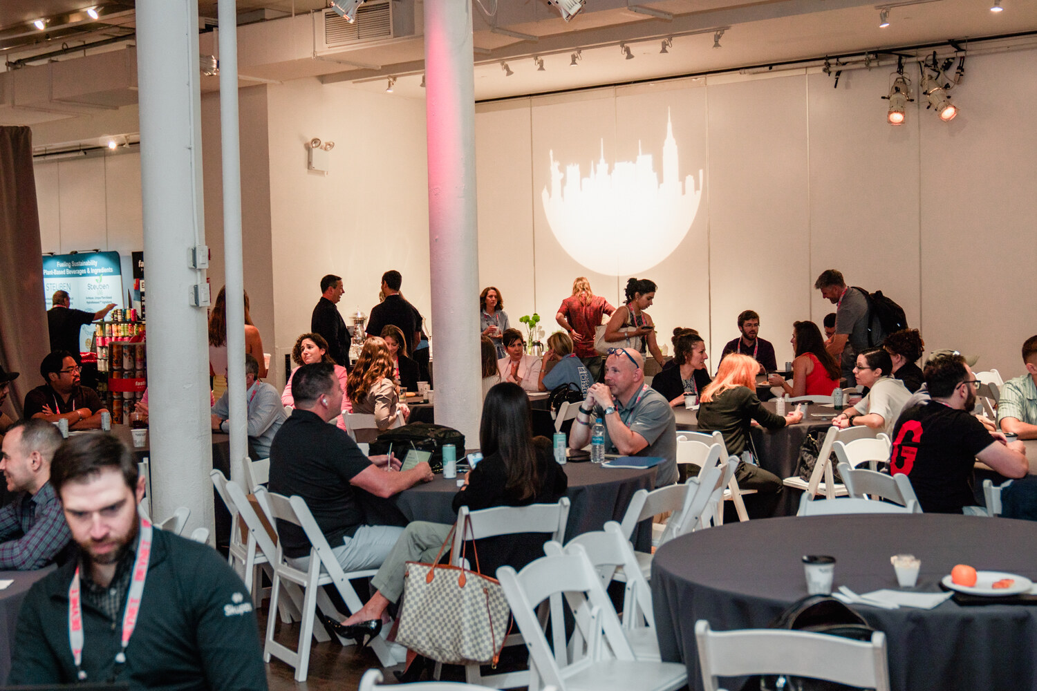Wide view of attendees seated at round gray tables in a loft room with columns and a New York skyline graphic on the wall.