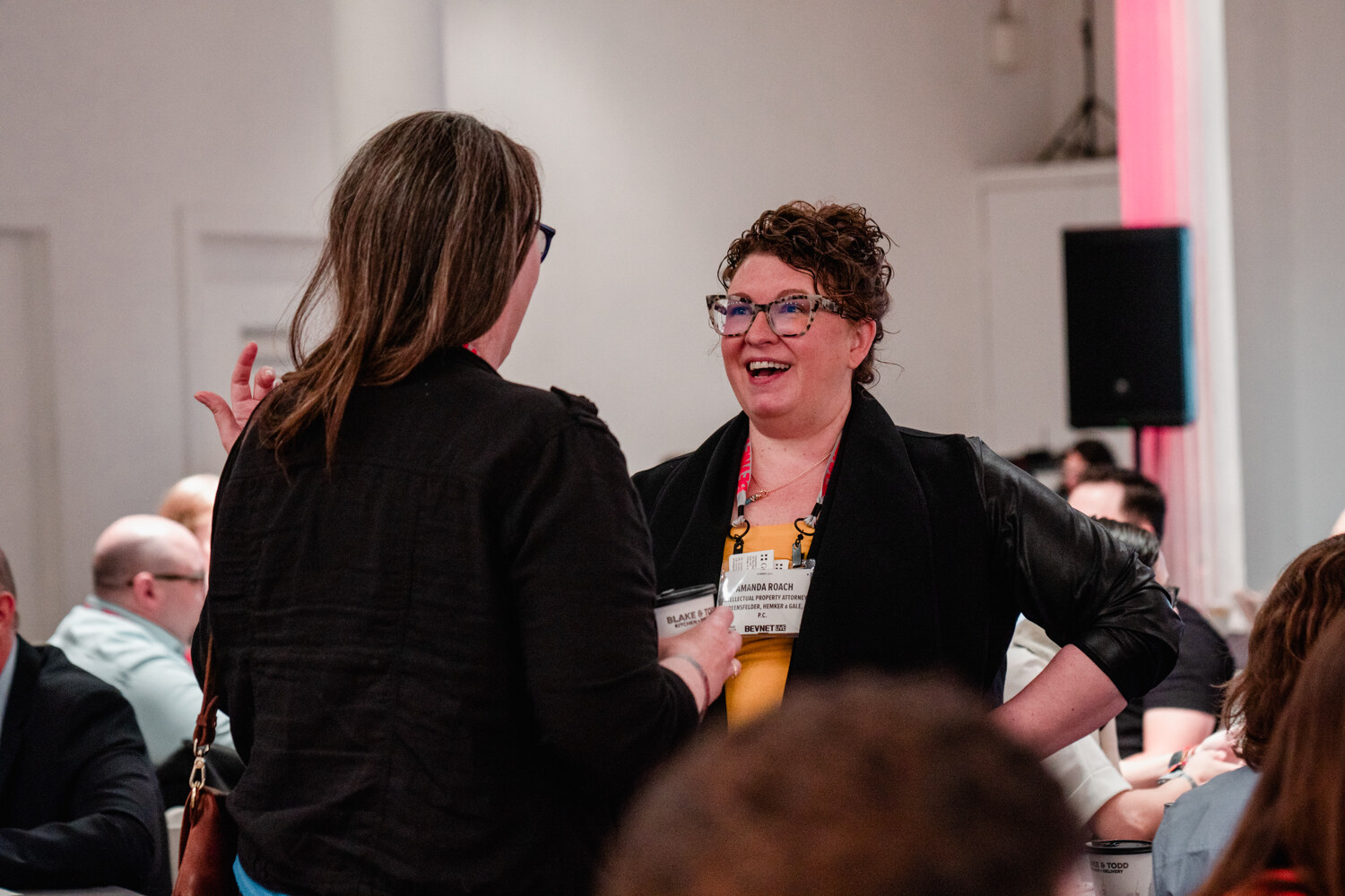 A woman with curly hair, glasses, and a yellow name badge laughs while talking with a woman in black who has her back to the camera.