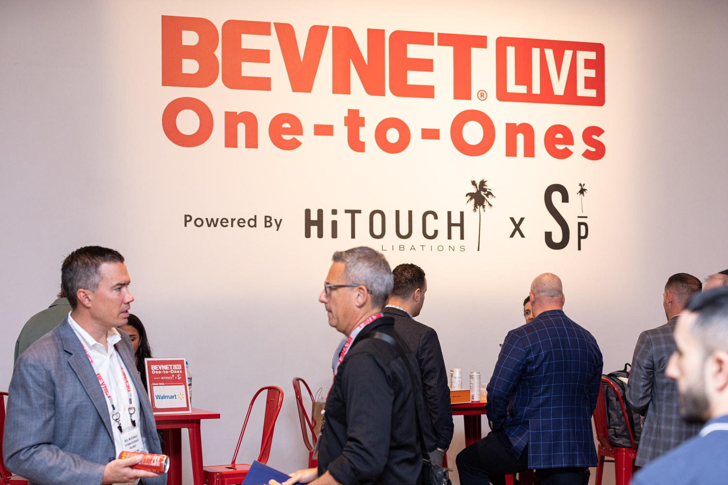 Several men in jackets and patterned shirts stand and talk in front of a white wall with the BevNET Live One-to-Ones sign and Hi-Touch Libations logo.