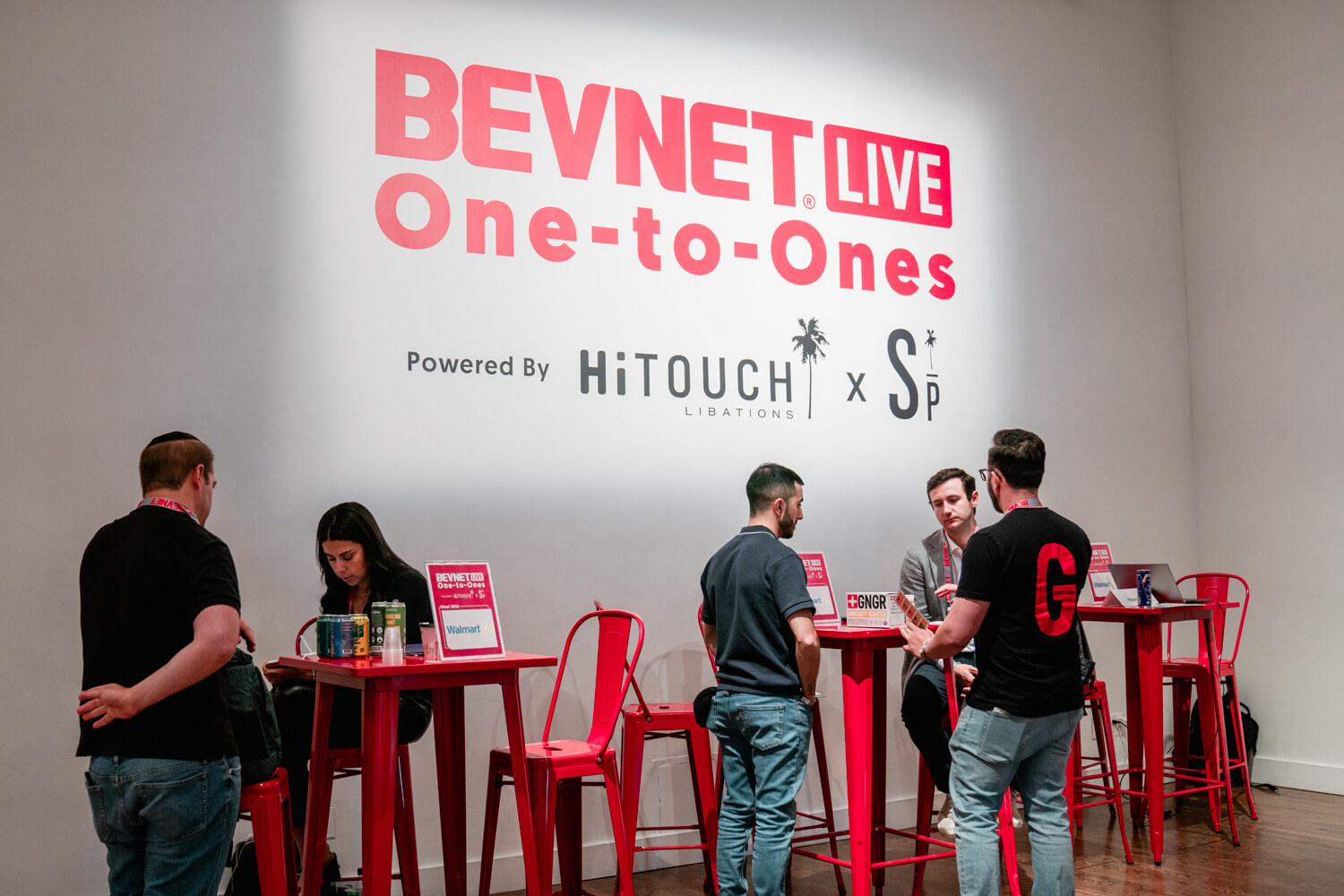Wide view of people sitting and standing at red high tables in front of a lit wall sign reading BevNET Live One-to-Ones powered by Hi-Touch Libations.