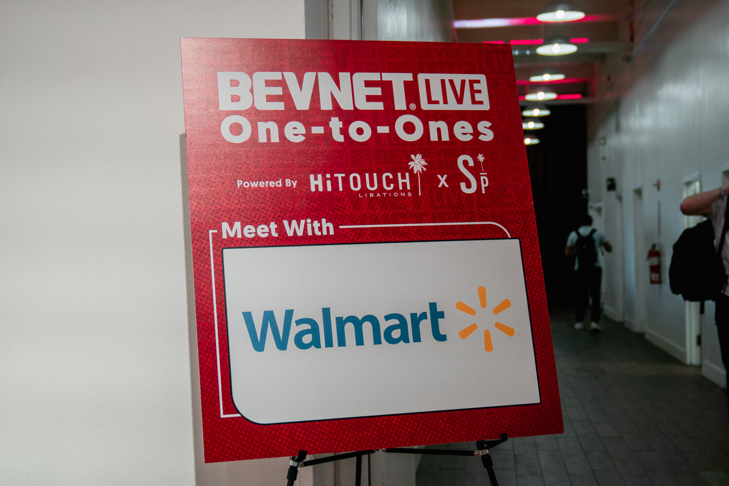A red easel sign reading BevNET Live One-to-Ones, Meet With, and a Walmart logo stands in a hallway.