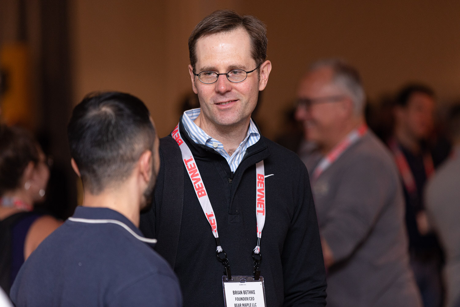 A smiling man with glasses in a black pullover with a BevNET lanyard stands in a busy room with other attendees blurred behind him.
